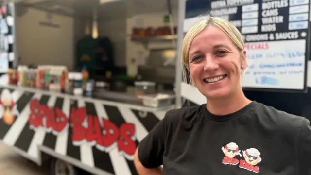 A woman stands in front of a black and white striped food truck named "Spud Buds" with the name in red letters. A menu board is visible in the background.