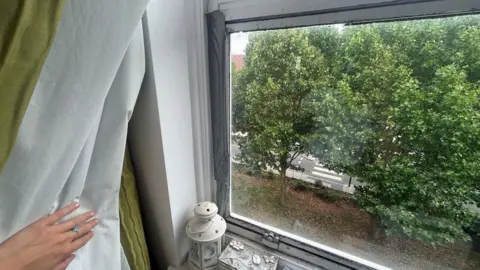 Picture showing a metal-framed window with silver duct tape attached to the left hand side. You can also see a hand pulling back a curtain and out of the window are trees and a zebra crossing. 