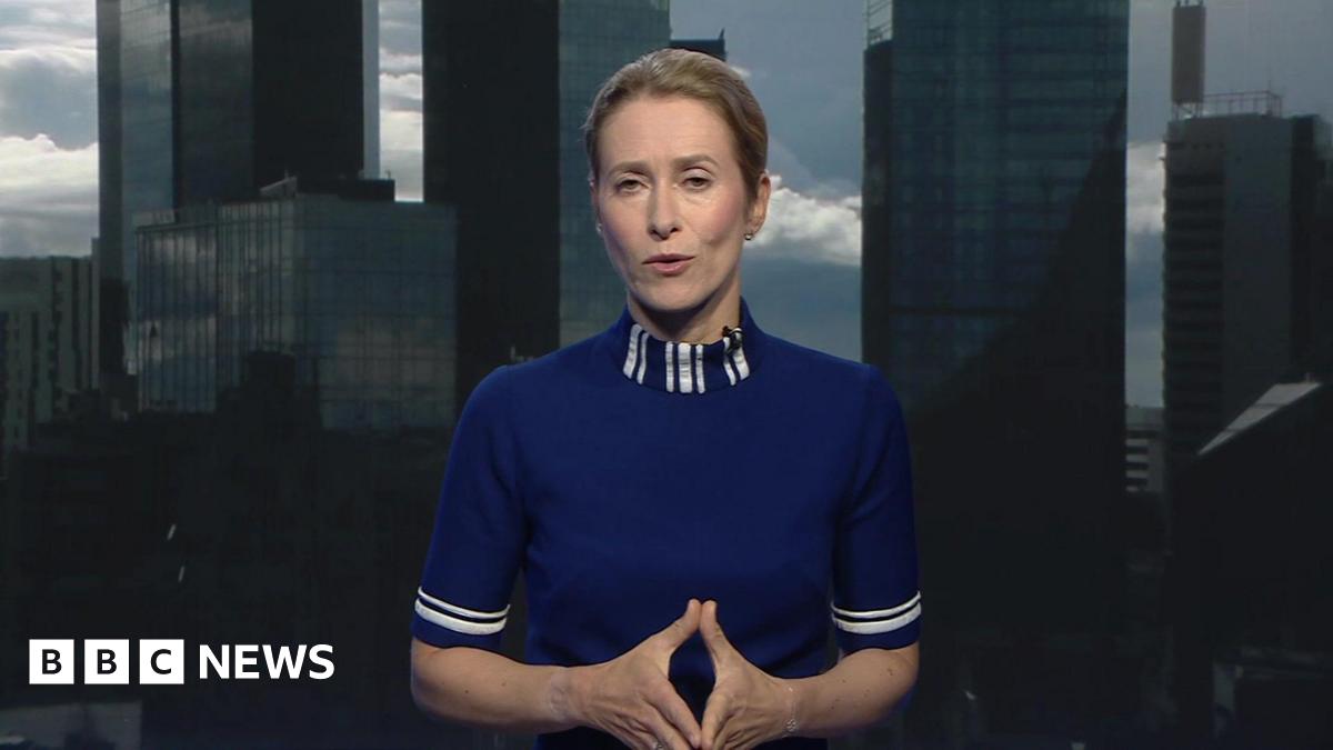 A blonde woman in a high-necked blue top is speaking to a camera, with a microphone clipped to her collar. Her hair is tied back and she is gesturing with her hands pressed together.