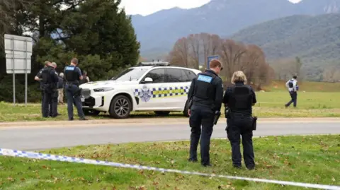 EPA/Shutterstock Police talking on the side of a country road, with a Highway Patrol vehicle in the background and police tape in the foreground. The landscape is green and mountainous.