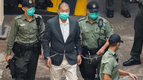 Reuters Lai, dressed in a gray suit and beige pants, walks handcuffed and flanked by three police officers to a police van