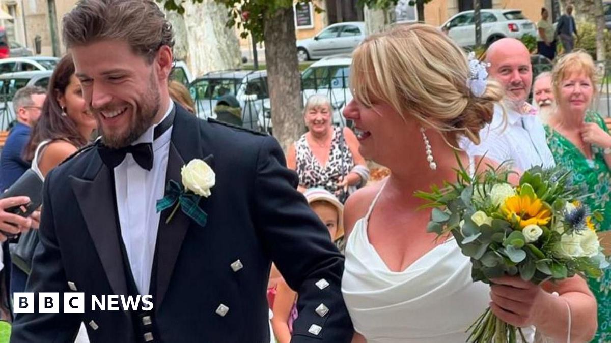 A white house with windows with brown shutters and a green lawn, with trees and hills in the background