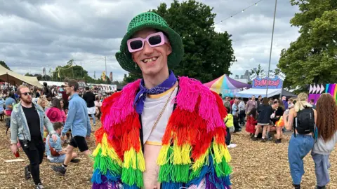 Joey Knock A man smiles at the camera at a music festival. He is wearing a green crocheted bowler hat, pink thick-rimmed sunglasses and a purple bandana around his neck. He's also wearing a mesh cropped tanktop and a rainbow coloured jacket covered in different coloured tassels. People can be seen in the background enjoying the festival.
