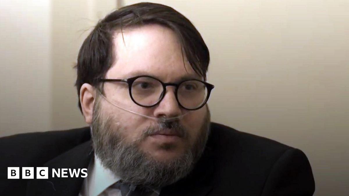 A bearded man with an oxygen tube in his nose. He is wearing glasses and a dark suit