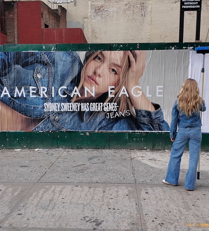 Woman in denim jeans stands in front of an American Eagle billboard featuring Sydney Sweeney advertising jeans.
