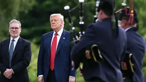 AFP via Getty Images Sir Keir Starmer and President Donald Trump are standing side by side in front of two male bagpipers, who have their backs turned to us. Sir Keir is in a dark blue suit with white shirt and blue tie. President Trump is in a dark blue suit with white shirt and bright red tie. They are all standing in front of green trees.