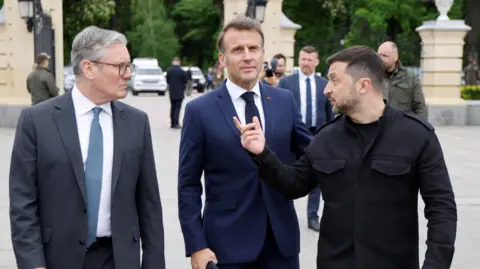 Reuters Volodomyr Zelensky, Emmanuel Macron and Sir Keir Starmer walk side-by-side. Macron and Starmer are wearing suits, while Zelensky wears a black utilitarian jacket. They are walking in front of a large stone and wrought-iron gate, behind which stand security, several cars, and a row of trees.