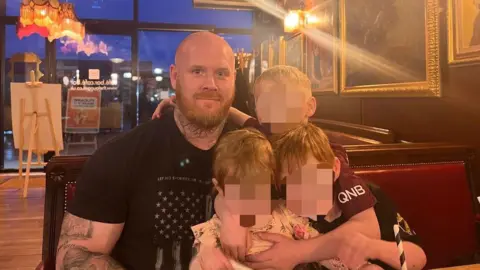 Lancashire Police Lenny Scott, who has a bald head and a ginger beard and wears a black t-shirt, poses with his children in a restaurant.