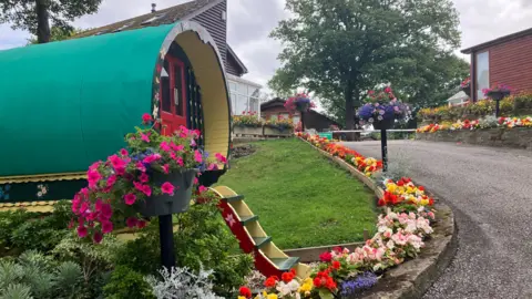 A recreation of an old gypsy caravan sits at the entrance to the caravan park. There are colourful flower beds leading up the roadside from the main entrance