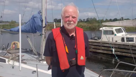 BBC A man with short grey hair and a grey beard, standing on a boat. He is wearing a navy t-shirt and a red life jacket. The boat is sitting on Lough Neagh, which is visible in the background. 