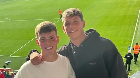 Nathan Black Two men stand smiling at the camera. The man on the left has on a cream T-shirt and has short brown hair. The man on the right also has short brown hair, he wears a black hoodie. Behind them is a green football pitch. 