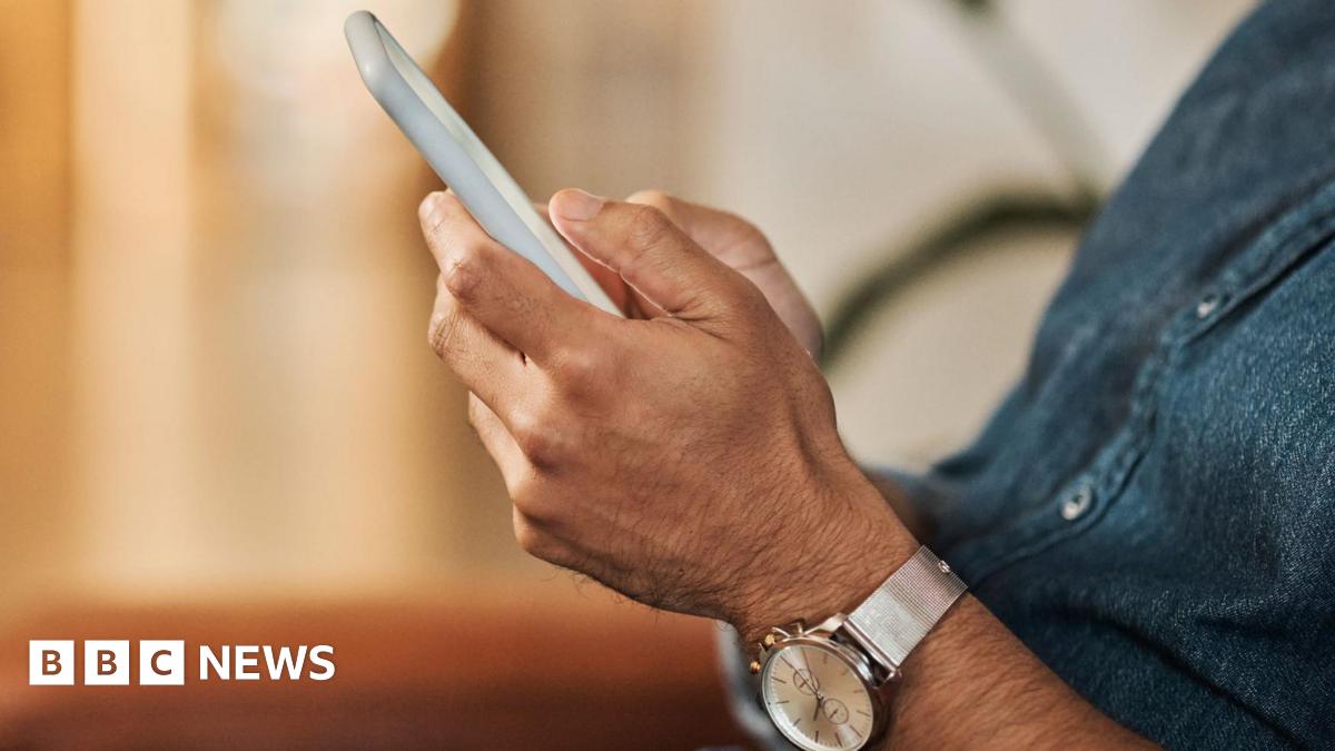 A man wearing a silver watch and denim shirt holds a white smartphone in his hands, his face is not visible nor is his screen