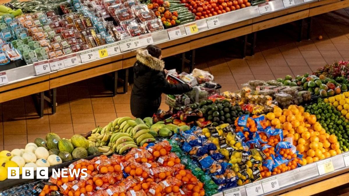 A woman in a dark coat and seen from above pushes a shopping cart through the produce section of a grocery store. On either side of her are piles of brightly coloured produce.