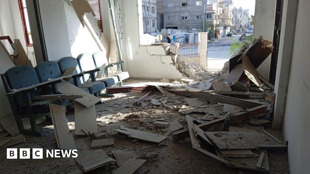 A badly damaged wall of an office, debris is scattered inside it and dust covers a bank of witting room chairs