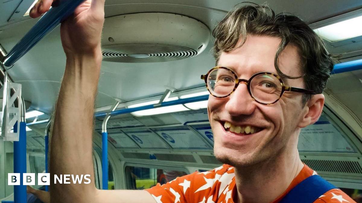 A man on the London underground smiles at the camera. He's holding his right arm up to hold onto the handrails and the photo looks like it was taken candidly. He's wearing an orange t-shirt with white stars and round tortoise-shell glasses.