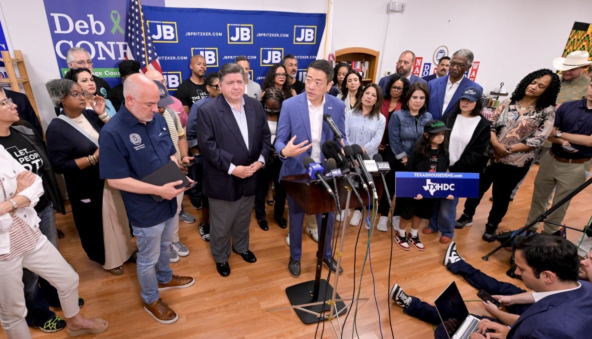 A group of people stand in a room in front of an array of microphones. A man in a suit jacket is talking into the microphones. 