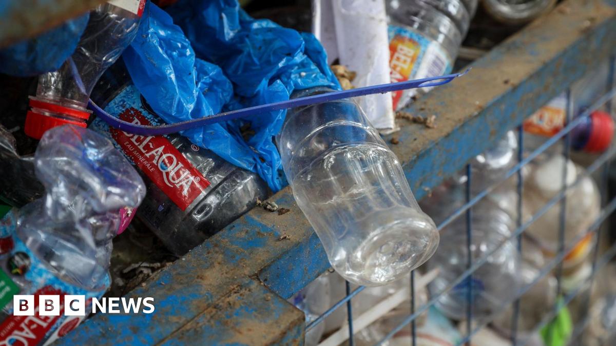 A blue metal gridded bin is stuffed full of plastic bottles of different colours. At the top of the bin a clear plastic bottle hangs over the side of the bin, on top of it is blue plastic and a blue cable tie