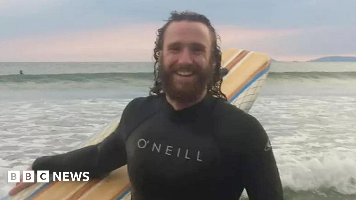 Alexander Barrett wearing a wet suit and holding a surf board while standing in front of the sea. He is smiling and has long brown hair and a brown beard.