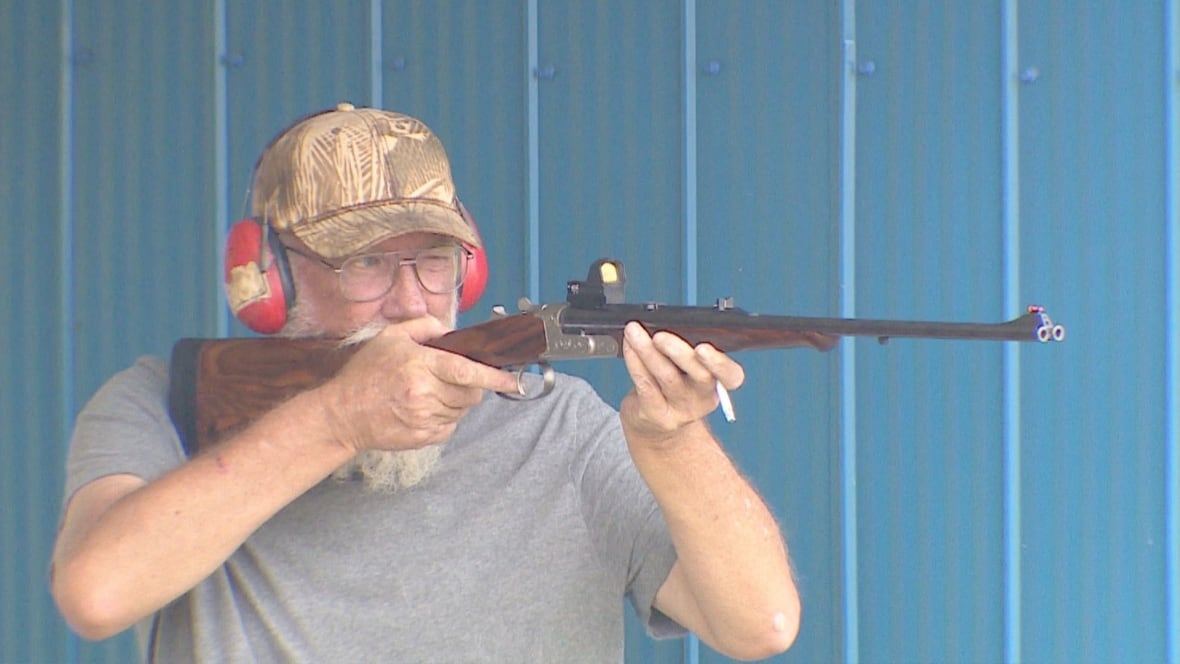 A man looks down the scope of a gun in front of a blue backdrop.