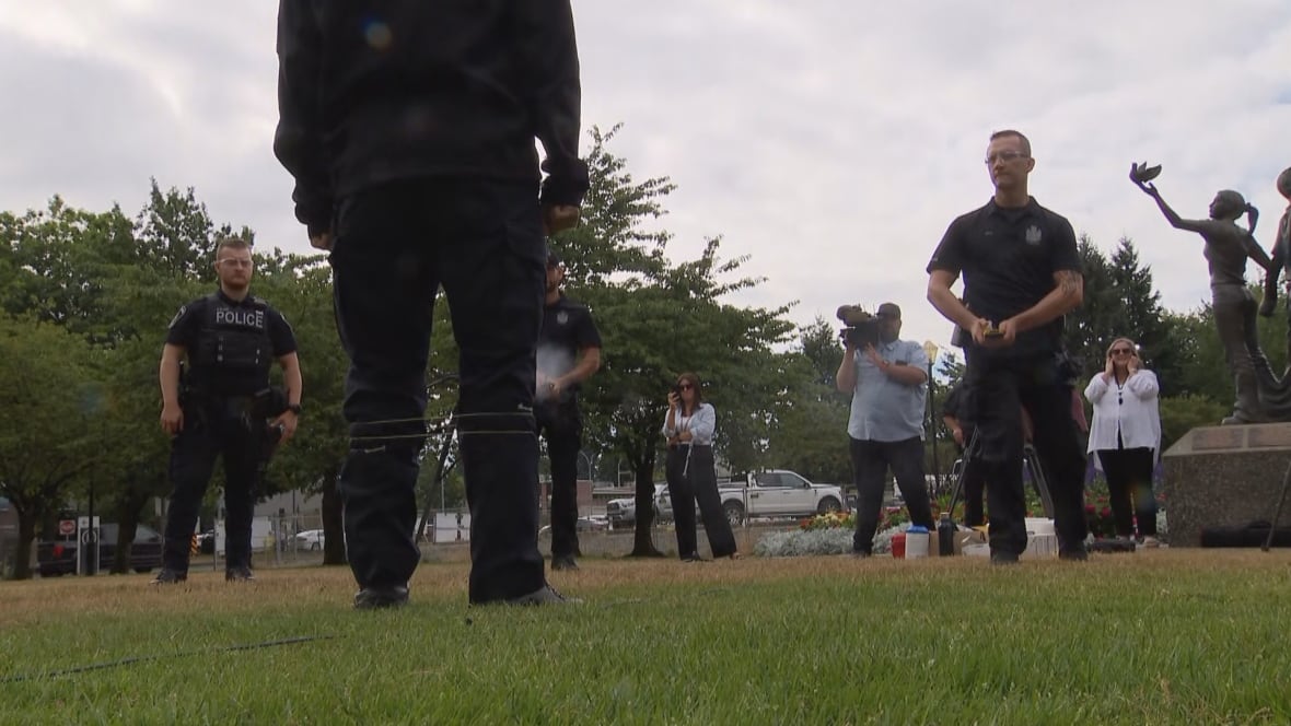 Police officers stand around. One of them is handling a device. Television cameras are recording.