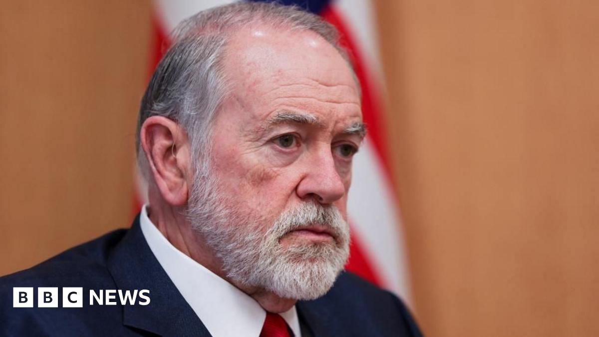 Mike Huckabee, who has grey hair and a short grey beard and moustache, looks on during an interview at his office at the US embassy in Jerusalem. An American flag is behind him.