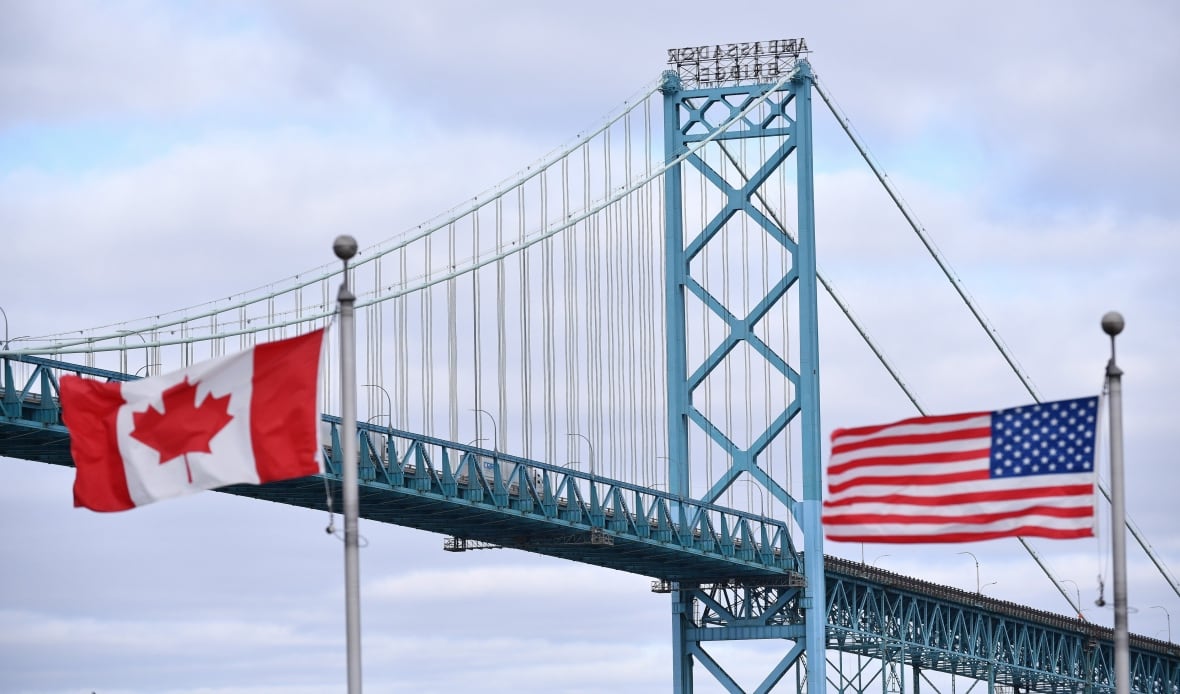 Canadian and American flags fly near the Ambassador Bridge at the Canada-USA border crossing in Windsor, Ont. On the left is the red and white Canadian flag, with a maple leaf in the centre and red vertical bands on the left and right sides. On the right is the American flag, with its red and white horizontal stripes and white stars on a blue background. The bridge spans across the image in the background against an overcast cloudy sky.