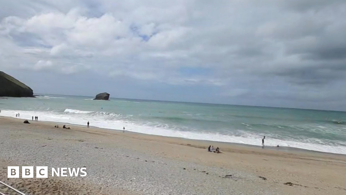The picture shows Portreath beach which has a mix of sand and pebbles. Several people are visible, some standing near the water, others on the sand. The ocean has gentle waves, and a large rock formation is visible in the water near the horizon.