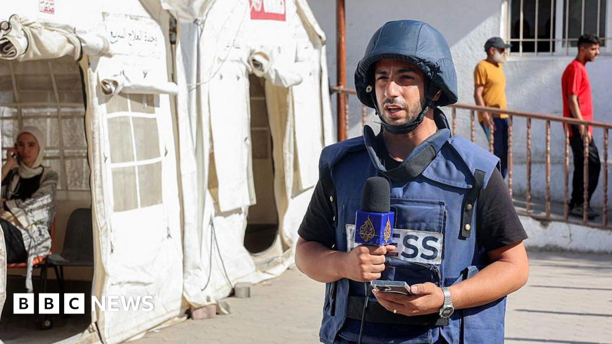 Anas al-Sharif stands next to a tent wearing a dark blue helmet and vest with "press" written on it, while he speaks into a microphone during a broadcast.