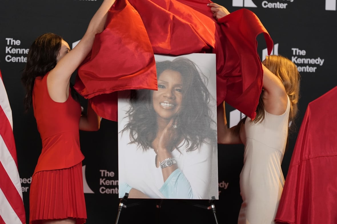 Two women in dresses on a state take drapes off of a portrait of another woman.
