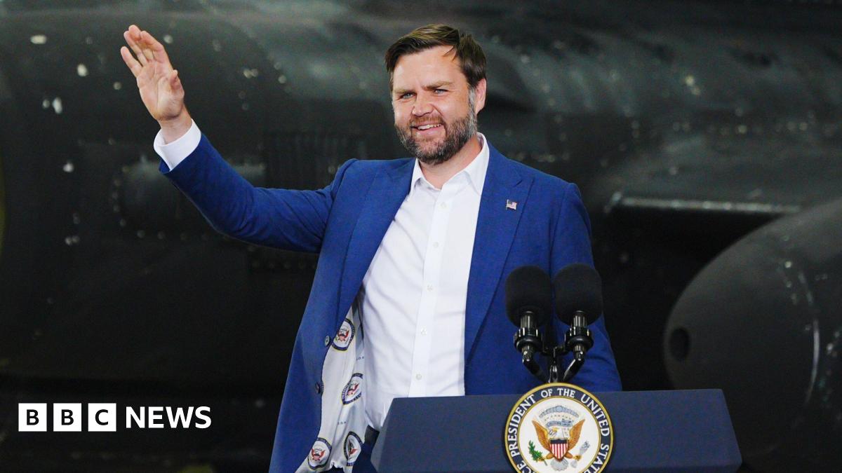 JD Vance stands waving at a lectern in front of a black US military plane in a hangar at RAF Fairford - he wears a blue suit jacket and white shire. The lectern has two microphones and the emblem of the US Vice President's Office