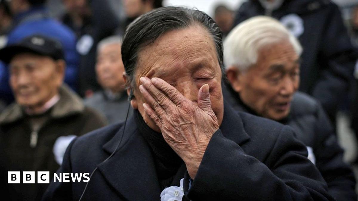 An elderly man wearing a suit with an ear piece in his ear wipes tears off his face. Behind him more elderly men can be seen, slightly blurred