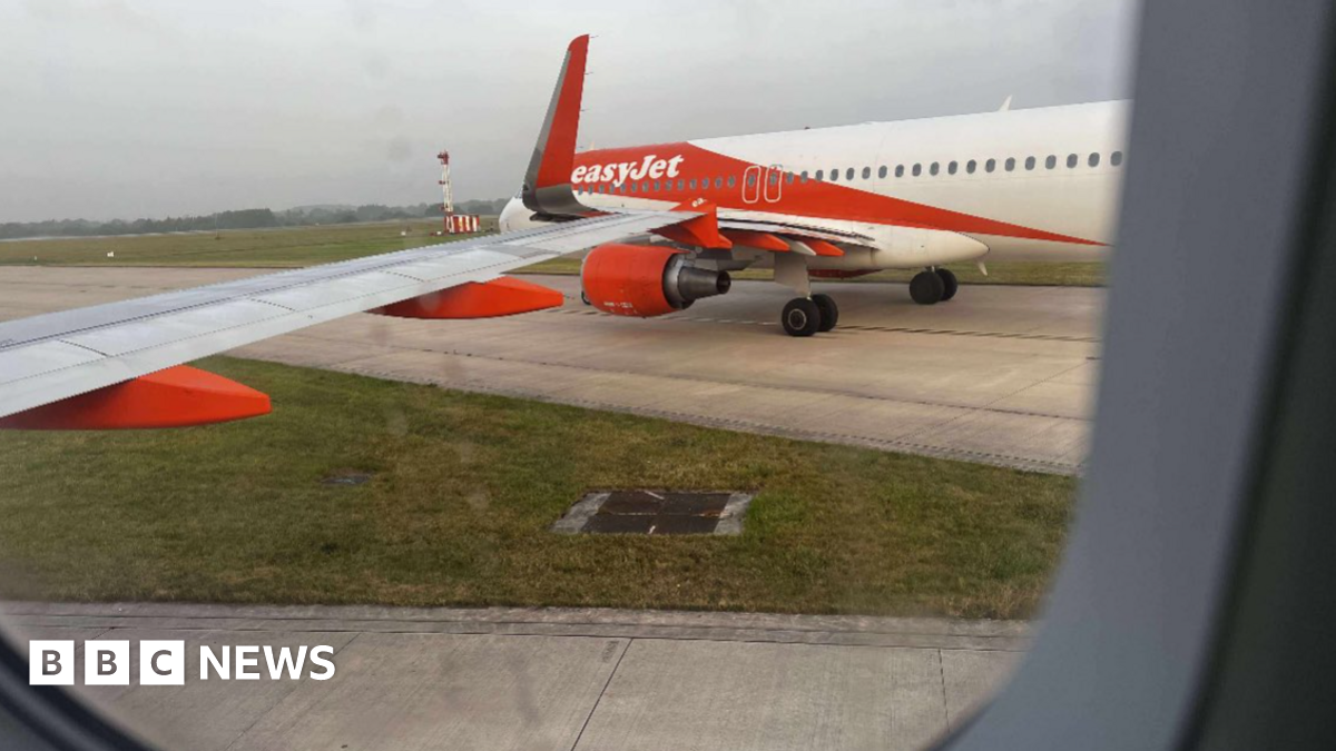 Image taken from a plane window shows the other orange and white liveried EasyJet plane on another taxiway. Both taxiways are joining together at a junction where their wings have clipped.