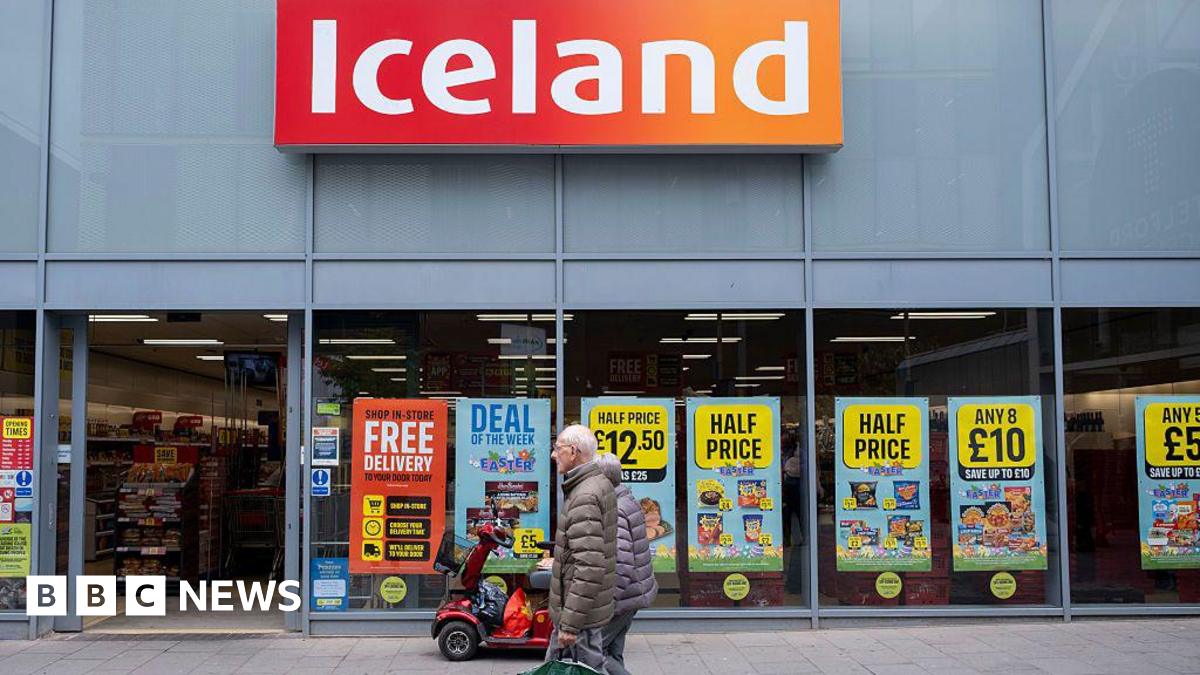 An elderly couple walk outside an Iceland store with lots of posters advertising discounts in the window