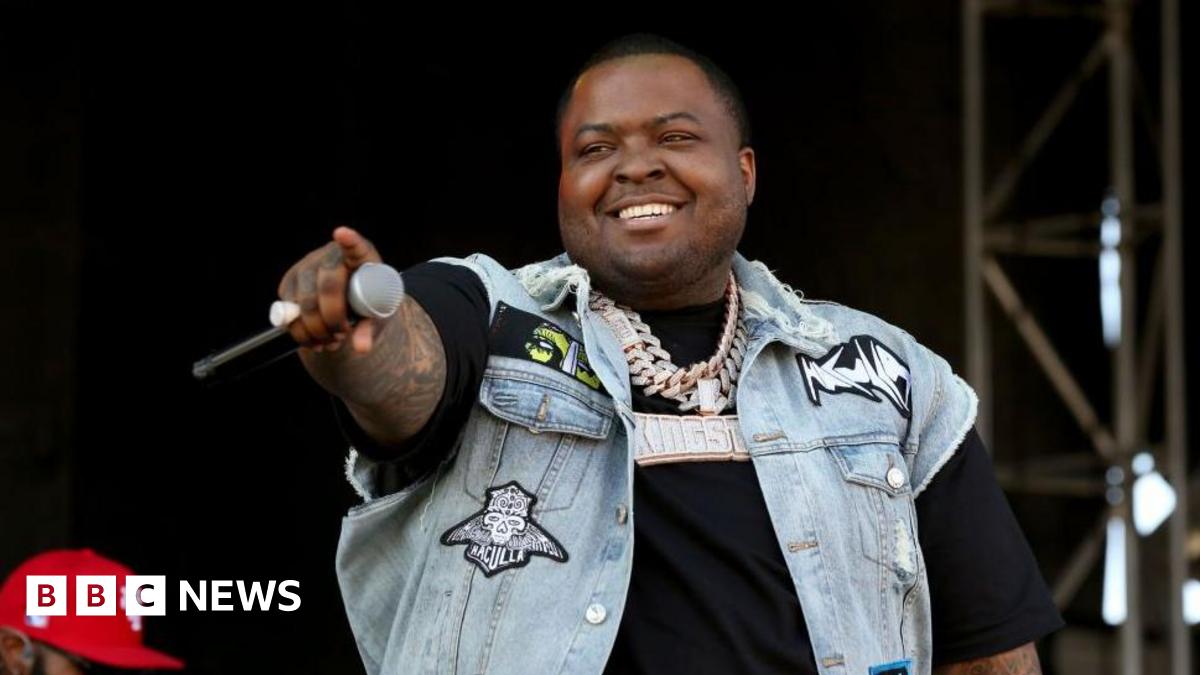 Rapper Sean Kingston is seen on stage holding a microphone and pointing out into the audience. He is smiling and wearing a black T-shirt with a jean vest over it.