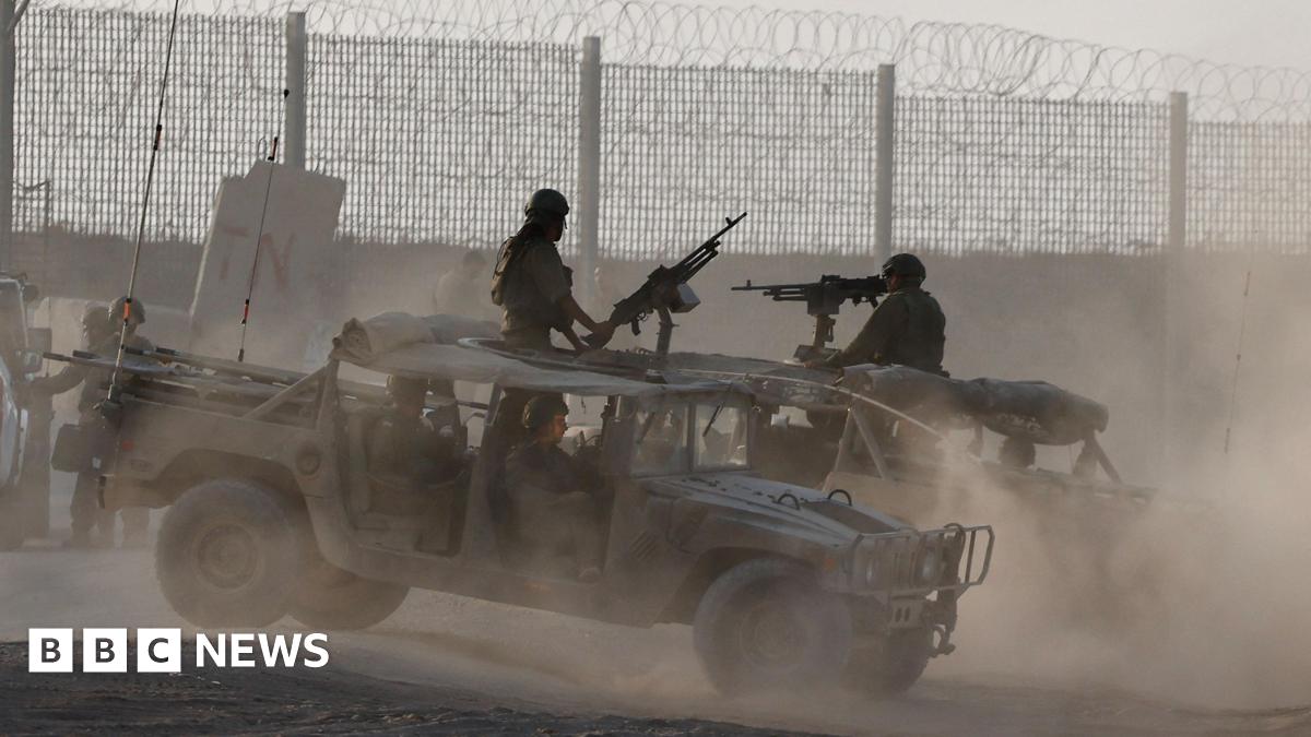 Members of the Israel Defence Force stand in two open-topped military vehicles near a fenced border, which is topped with barbed wire. Each vehicle has a large gun extending from its roof, manned by a person in a helmet and camoflague uniform.