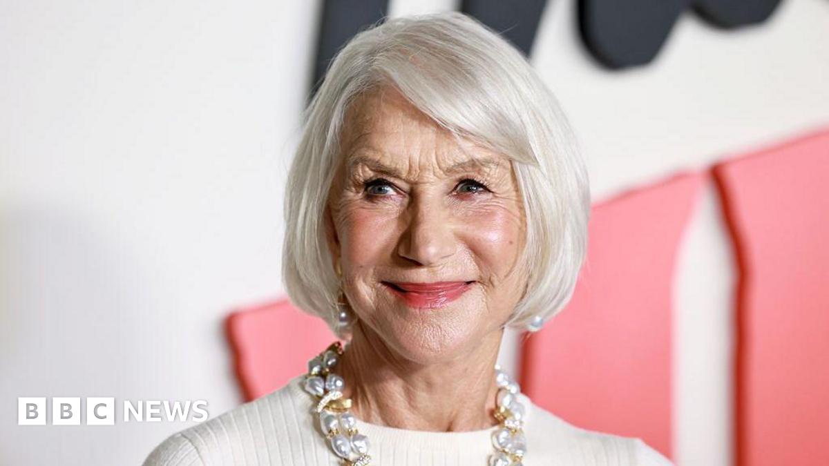 An older woman with white hair cut in a chin-length bob is looking to the right of the camera. She is wearing pearl earrings and a chunky gold and pearl necklace, with a high necked white top.