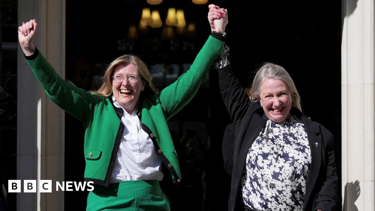 Two women celebrating outside the Supreme Court. The woman on the left is wearing a green suit with the jacket open over a white shirt. She has her hands in the air. Her left arm his being raised by the woman on the right, who has her right arm in the air. She is wearing a black jacket, which is open, over a blue and white floral patterned top.