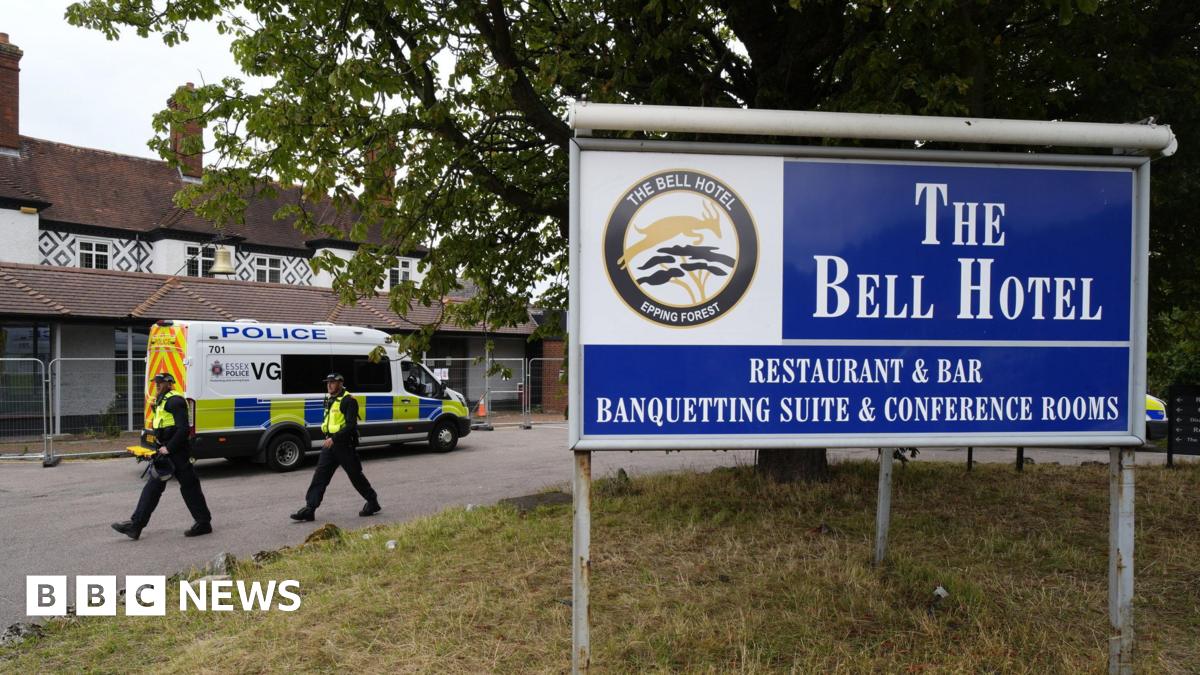 Two police officers walking next to the hotel, which is surrounded by a thin metal fence. A police van has also parked next to the building. In the foreground is a large blue sign which reads 'The Bell Hotel'.