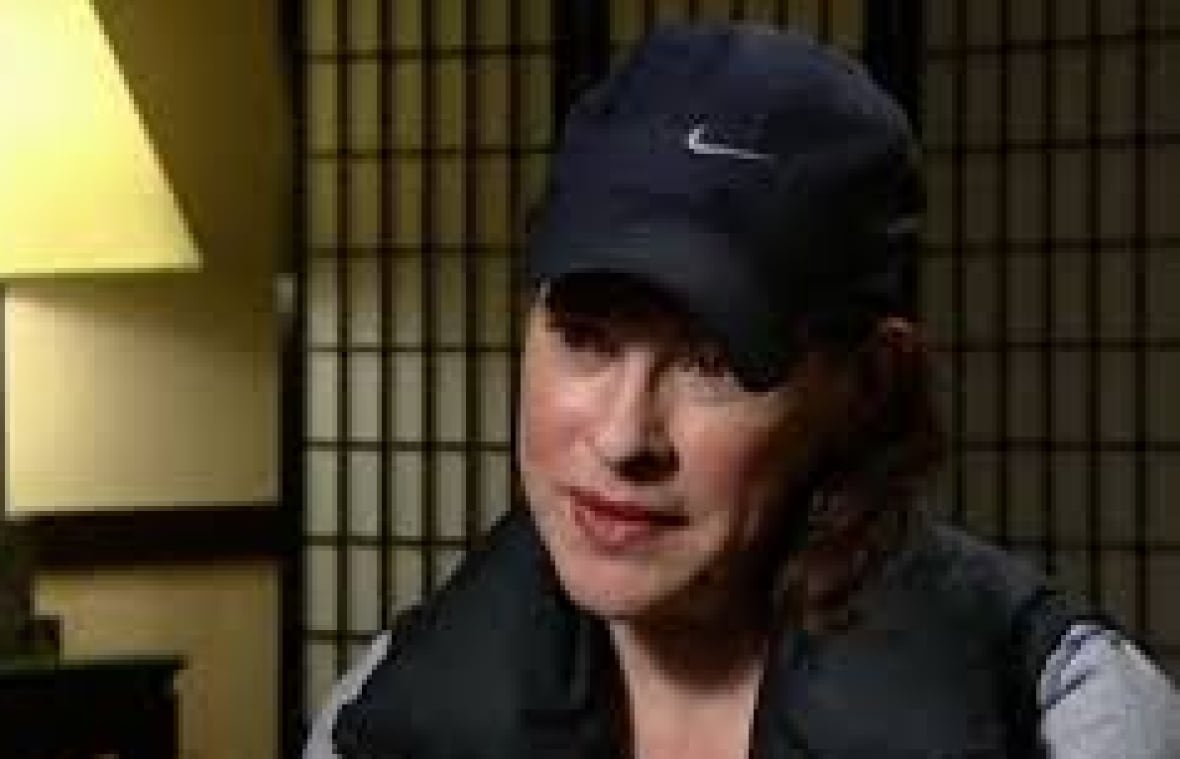 A woman wearing a baseball cap speaks in a room in front of a foldable screen.