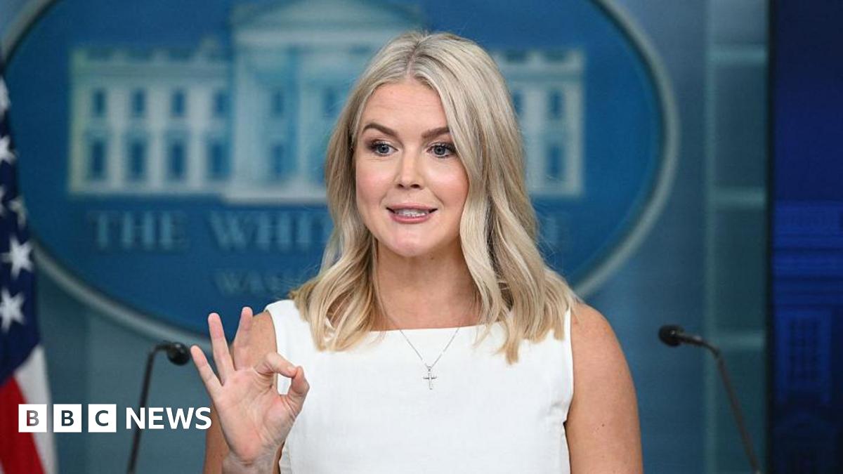 Karoline Leavitt, White House Press Secretary, delivers remarks during the daily briefing held in the Brady Briefing Room at the White House. Standing behind a lectern, she is dressed in white, maintains eye contact with the audience and gestures with her right hand in an "OK" sign.