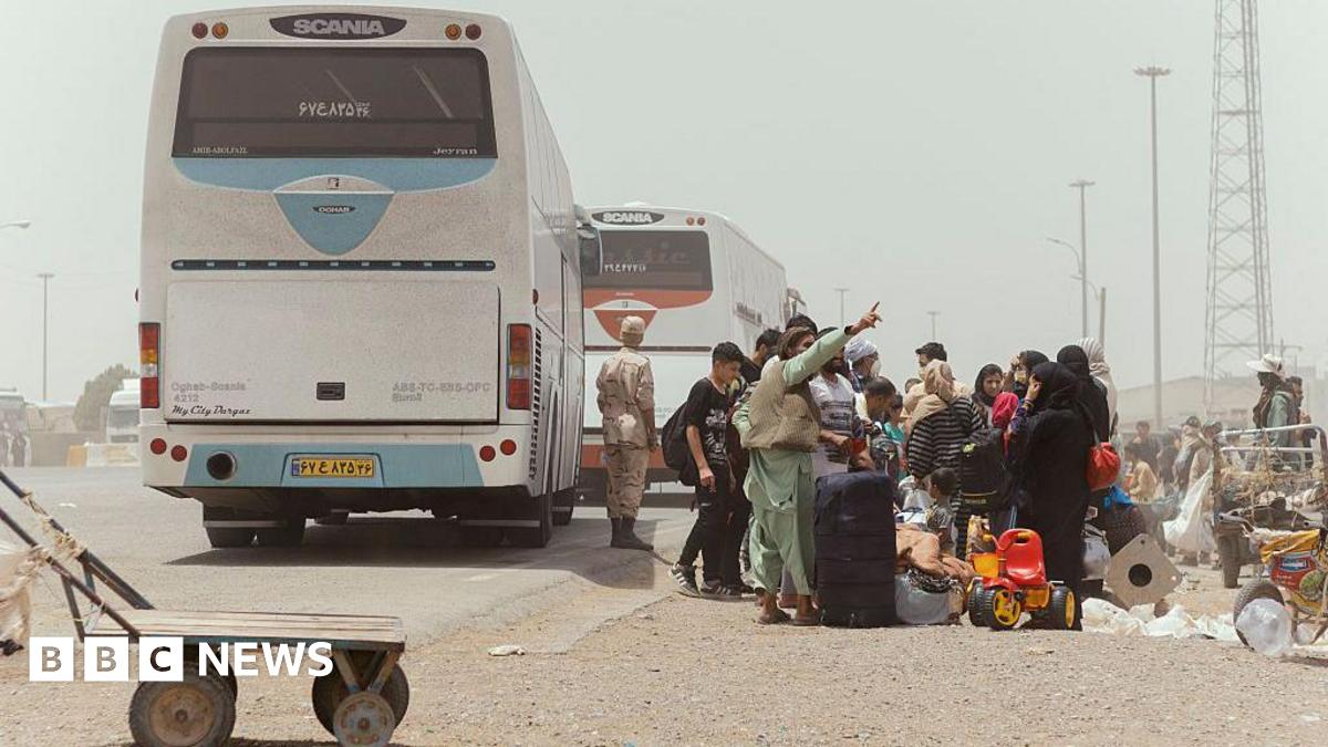 Afghan deportees crowding beside a parked bus, on a dusty road. They are carrying bags and suitcases.