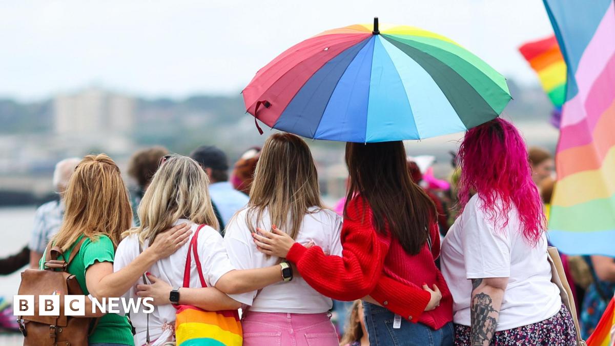 A group of people at Liverpool Pride are facing away from the camera and standing under a rainbow umbrella. They are wearing colourful clothes, and one has a rainbow bag while another has pink hair. A rainbow flag is nearby.