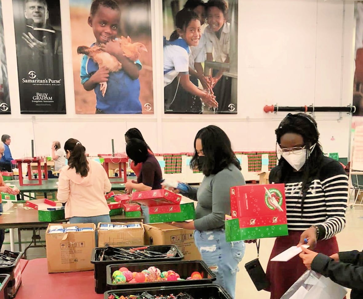 Young volunteers in face masks work in a large gymnasium filling red and green boxes, drawing from bins filled with small toys and items on tables before them.
