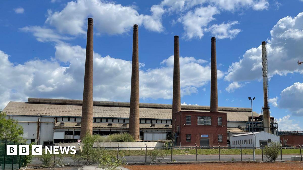 A factory with five tall narrow chimneys rising from the ground. The sky is blue with some white clouds floating above.