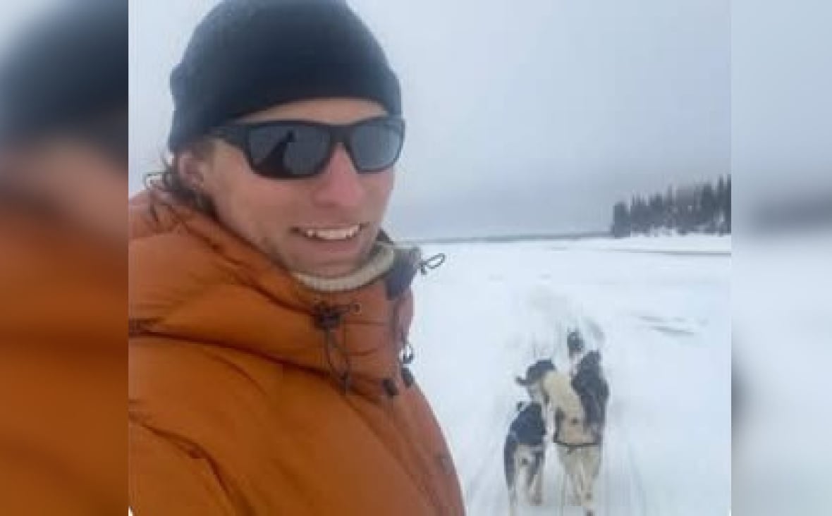 A man wearing a black tuque and orange parka and black sunglasses looks towards the camera. It's snowy outside. There are dogs in the background.