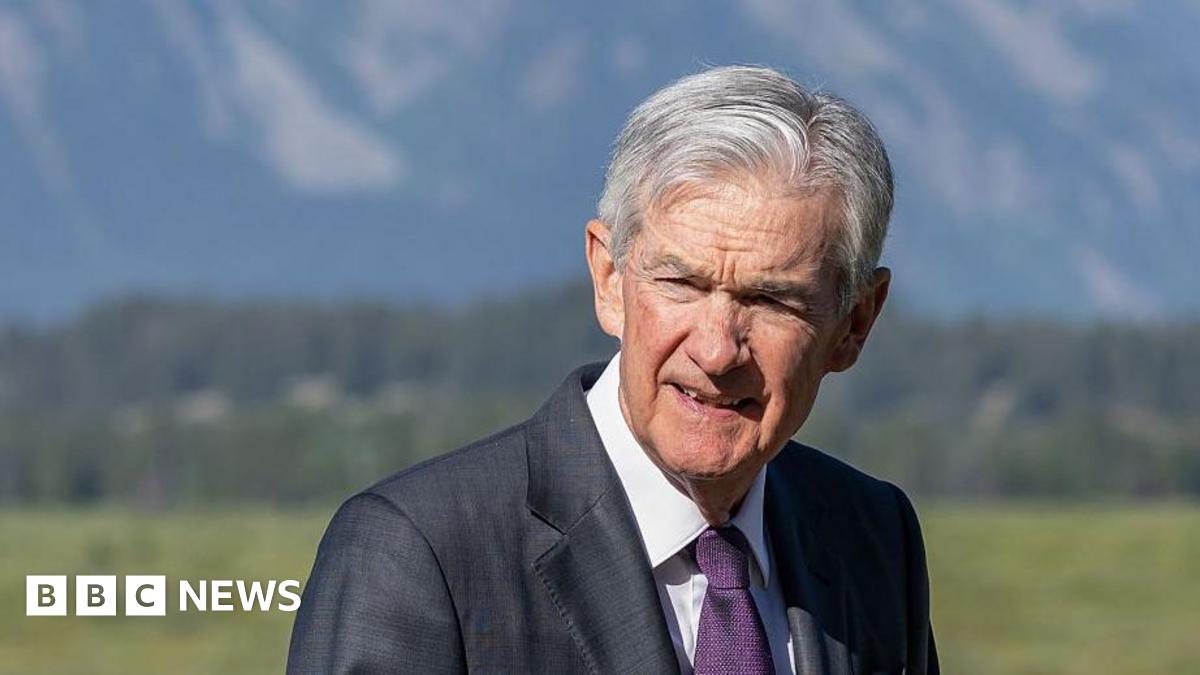 Jerome Powell squinting in the sun, wearing suit and tie outdoors in Jackson Hole with trees and mountainsides in background