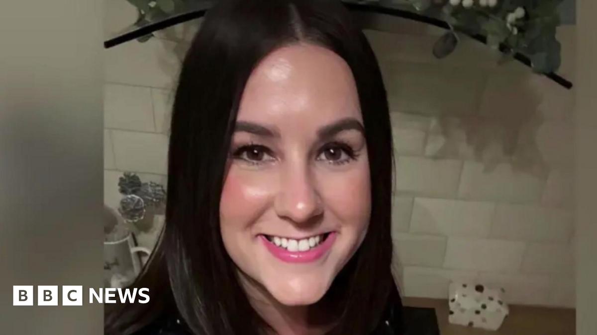 A smiling Lucy Connolly looking directly down the camera. She has shoulder length, dark brown hair and brown eyes. She is seen indoors, in what appears to be a kitchen, with a white tiled splashback and work surface behind her. The edge of a hob extractor fan can also be seen behind her.