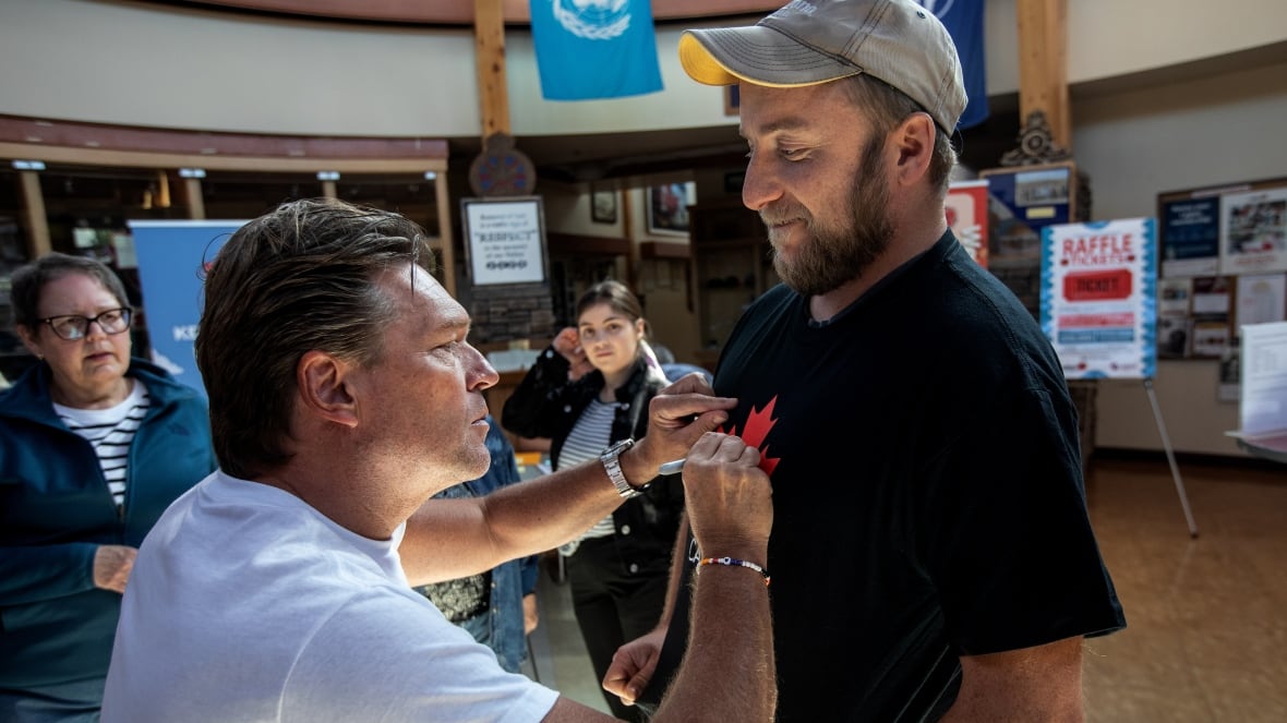 A white man, with slicked back greying hair, is wearing a white T-shirt. He is signing the T-shirt of another white man, who has a beard and is wearing a ball cap. They are inside a room; two women watch them.