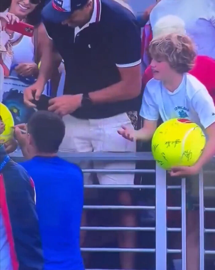 Crowd at US Open tennis event with fans interacting and a boy holding a signed yellow ball near a railing.