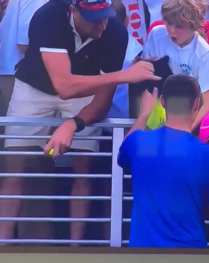 Polish CEO reaching over railing to grab a hat during US Open, with surprised child and other spectators nearby.